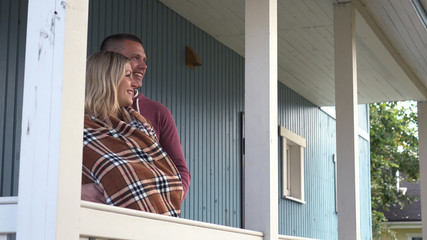 Happy young man hugging his pregnant girlfriend in a blanket standing outdoors. Country house blue.