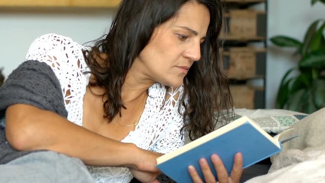Woman in casual clothes looks absorbed while reading book on the sofa
