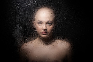 Portrait of a bald girl behind a wet glass on a black background.