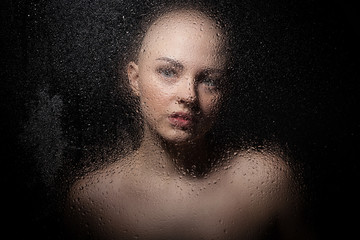 Portrait of a bald girl behind a wet glass on a black background.