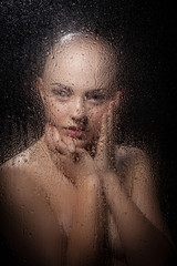 Portrait of a bald girl behind a wet glass on a black background.