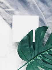 Closeup of white plain square box or package next to a big tropical leaf and grey scarf on white marble desk background. Copy space. Flat lay. Top view