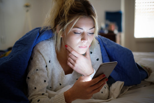 Disappointed sad woman holding mobile phone while laying on bed in a bedroom
