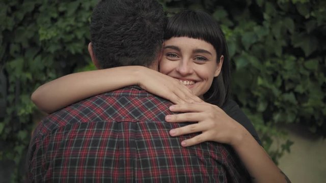 Sad and lonely man stands on street, appears from back his girlfriend or best friend or partner, beautiful young brunette, hugs and adores him in loving and tender hug, smiles into camera