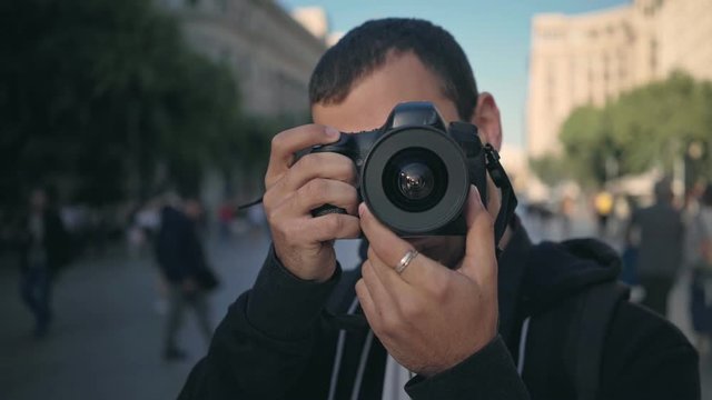 Young male tourist or photography student photographs into camera with big DSLR, clicks shutter to create memories. he smiles and laughs during summer vacation trip in europe