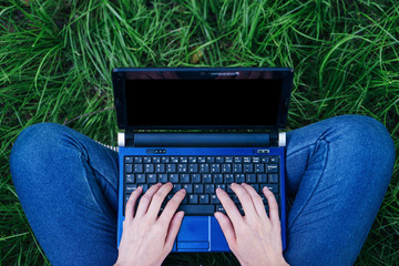 Top view of female hands typing on blank screen laptop computer while sitting on the grass. Freelance working concept.
