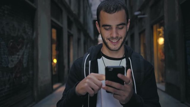 Attractive and handsome young man with beautiful smile and hipster millennial generation z looks, scrolls through social media feed on smartphone, giggles and smiles from news on applications