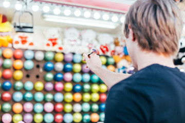Young man aims at wall of colourful air baloons in order to win teddy bear or plush toy prize for his girlfriend, during romantic date at amusement park or carnival