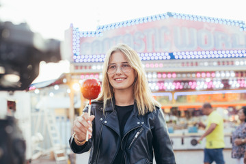 Attractive cute and adorable pretty blonde woman holds holiday sugar candy on stick, caramel toffee covered red apple in middle of carnvial festival fair in sunset light