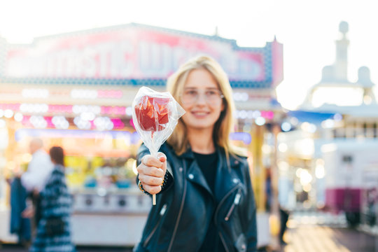Cute and pretty blonde happy woman in cool looking leather jacket poses for camera, shows and draws out hand holds toffee caramel sugar candy apple, at carnival fair