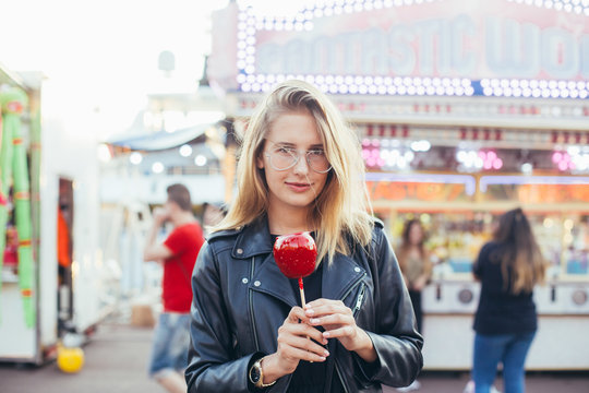 Attractive cute and adorable pretty blonde woman holds holiday sugar candy on stick, caramel toffee covered red apple in middle of carnvial festival fair in sunset light