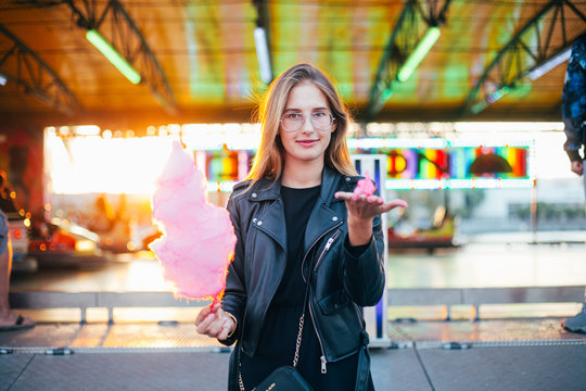 Adorable cute pretty woman stands in middle of amusement park, in front of ride with bright colours positive and cheerful, eats pink cotton candy floss, happy and optimistic