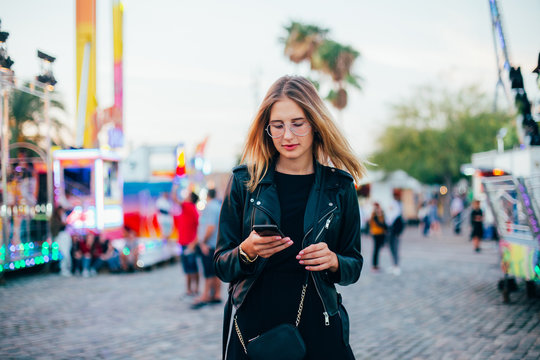 Beautiful woman chats on phone in middle of crows at local town amusement park or carnival fair, uses smartphone application to meet up or find friends, concept connectivity