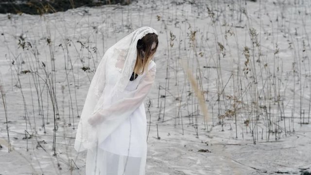 The young woman with scary make-up of dead bride for Halloween dressed in a white wedding gown going through the marsh with dry grass. 4K