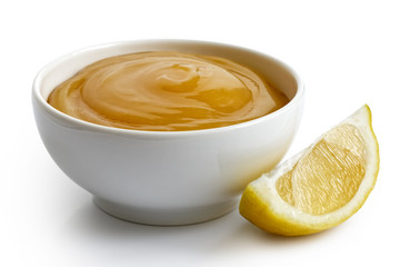 Lemon curd in white ceramic bowl next to lemon wedge isolated on white.