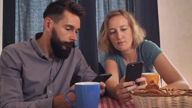 young happy couple using gadgets at home. Smiling girl holding smartphone showing boyfriend some funny picture. Boyfriend scrolling touch screen mobile indoors