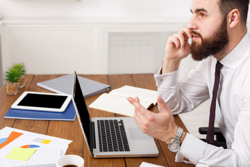 Serious businessman talking on cell phone with laptop in modern office