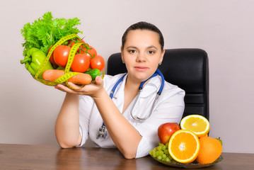 The doctor is a nutritionist in a white coat and a phonendoscope on his neck in his office at the table. The doctor keeps dietetic vegetables and fruits with vitamins.