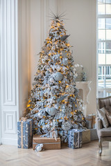 Украшение дома к Новому году. В доме стоит украшенная елка в белых и серебряных тонах с гирляндой. Под елкой лежат подарки