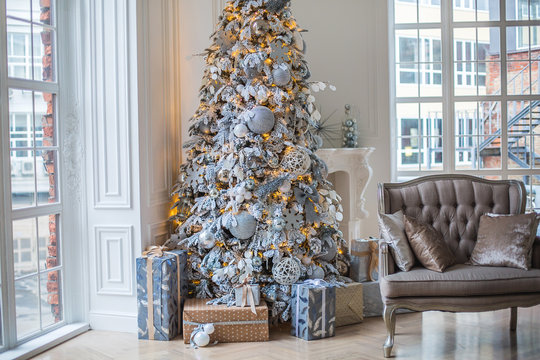 Украшение дома к Новому году. В доме стоит украшенная елка в белых и серебряных тонах с гирляндой. Под елкой лежат подарки