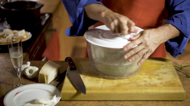 On the counter are a glass of champagne or proseco, garlic, fresh mozarella, and parmigiano reggiano as a chef brings a salad spinner over to his workstation and spins the water out of the fresh basil