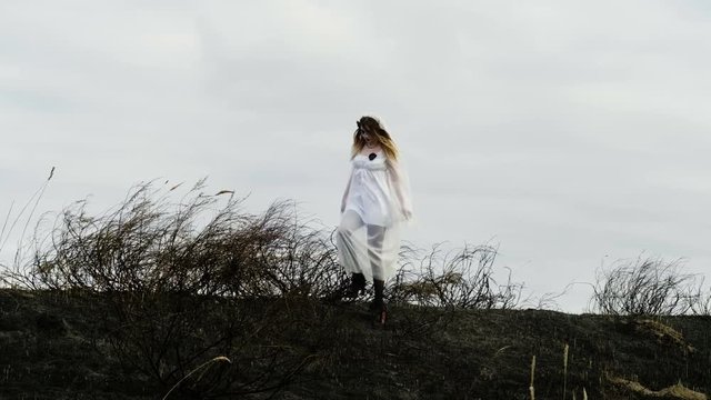 The young woman with spooky makeup for Halloween in a white bride dress walking through the field of dried grass on the background of the sky. 4K