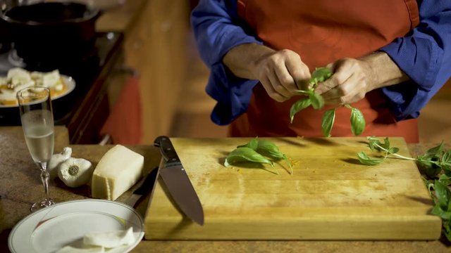 On the counter are a glass of champagne or proseco, garlic, fresh mozarella, and parmigiano reggiano as a chef brings pulls the leaves off of fresh branches of basil and puts them on his cutting board