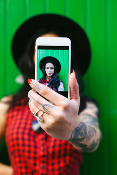 Portrait of a young alternative woman taking a selfie in front a green wall.