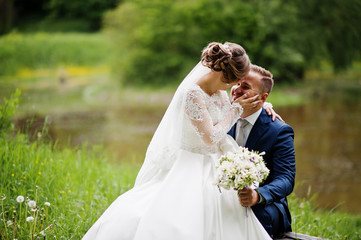 Fantastic bride sitting on a groom's lap in the meadow next to the lake on a wedding day.