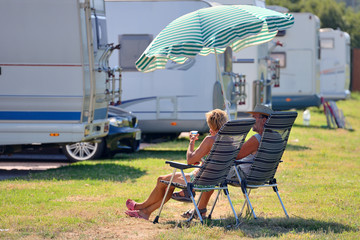 elderly couple is resting on chairs under a beach umbrella near a caravan
