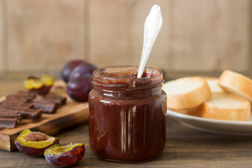 Plum chocolate jam in a glass jar, fresh plums and pieces of chocolate on a wooden surface.