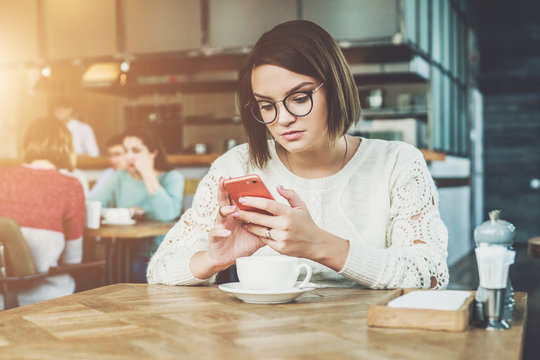 Young businesswoman is sitting in coffee shop at table and using smartphone, working. E-learning,online marketing,education.Hipster girl looking on screen of phone,checking email, blogging, chatting.