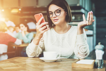 Young businesswoman is sitting in coffee shop at table and using smartphone, working. E-learning,online marketing,education. Hipster girl looking at phone screen in surprise, checking email, chatting.