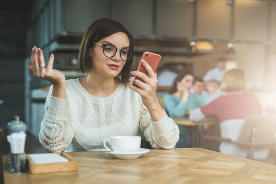 Young businesswoman is sitting in coffee shop at table and using smartphone, working. E-learning,online marketing,education. Hipster girl looking at phone screen in surprise, checking email, chatting.