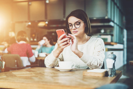 Young businesswoman in glasses and white sweater is sitting in cafe at table, using smartphone, working. E-learning,online marketing,education. Hipster girl looking on screen of phone, checking email.