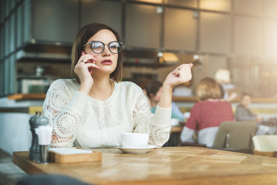 Young serious businesswoman in glasses sitting in cafe at wooden table and talking on cell phone. Telephone conversations. Hipster girl freelancer discusses working questions over phone. Girl calling.
