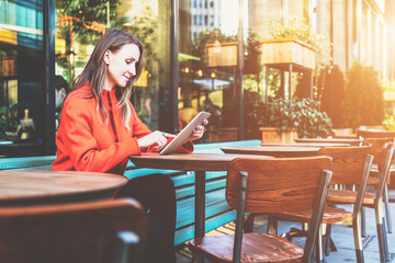 Side view. Young attractive woman in orange coat is sitting outside in cafe at table and uses tablet computer. Girl checking email, blogging, chatting, reading e-book. Social media, e-learning.