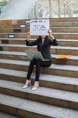 business woman unemployed from company sitting on the staircase and holding poster with text is looking for job, concept of unemployment problem