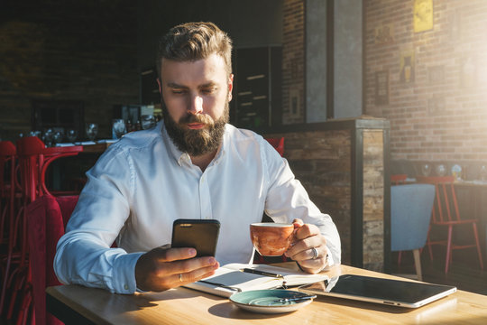 Young bearded businessman in white shirt is sitting in cafe at table, drinking tea and using smartphone. On desk is notebook and tablet computer.Online education, e-marketing, e-learning. Film effect.