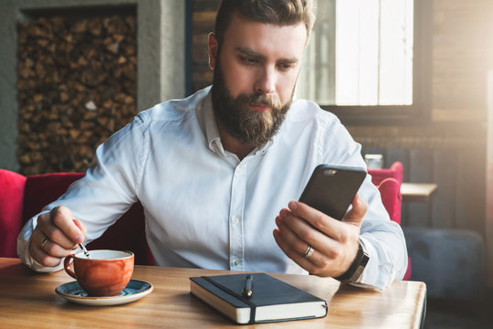 Young bearded businessman in white shirt is sitting in cafe at table,drinking tea and using smartphone.On desk is notebook.Man working, studying.Online education, e-marketing, e-learning. Film effect.