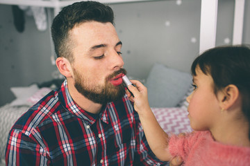 Cute little daughter and her handsome young dad in crowns are playing together in child's room. Girl is doing her dad a makeup