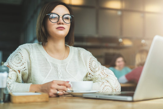 Young businesswoman in glasses is sitting in cafe at table in front of laptop. Student is studying online.Online education, marketing, e-learning. Social media, network. Instagram filter, film effect.