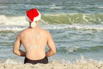 Man in Santa hats with the inscription New Year on the back on the beach. Back view