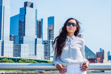 Fototapeta premium Young American Woman with long hair traveling in New York in summer, wearing white striped shirt, sunglasses, standing in business district with high buildings in Manhattan under sun, looking up..
