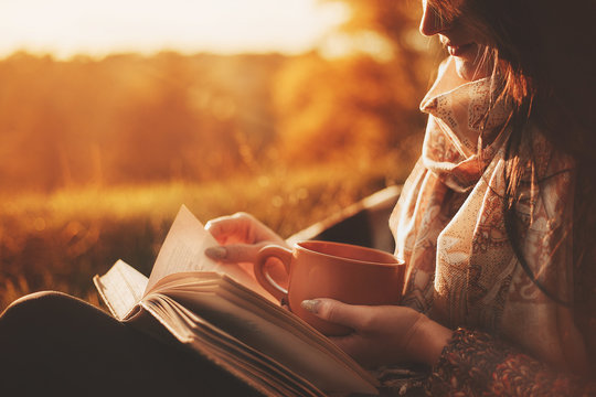 a woman sits near a tree in an autumn park and holds a book and a cup with a hot drink in her hands. Girl reading a book