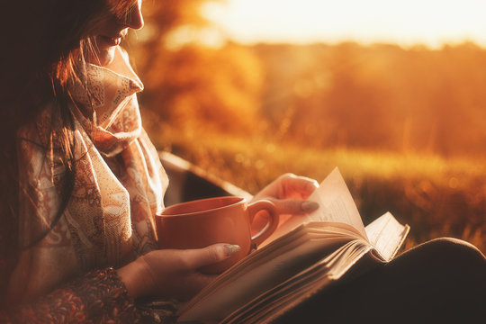 a woman sits near a tree in an autumn park and holds a book and a cup with a hot drink in her hands. Girl reading a book