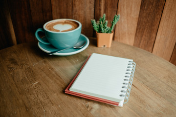 cup with coffee has a heart shape on top and diary notebook placed on wooden table . wood wall are background. image for business, coffee art, beverage, drink, food, art concept