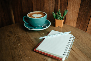 cup with coffee has a heart shape on top and diary notebook with pen on top placed on wooden table . wood wall are background. image for business, coffee art, beverage, drink, food, art concept