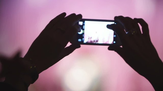 Closeup shot of hands of person taking picture of pop star or band performing on stage, crowd applauding, illuminated scene