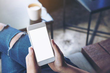 Mockup image of woman's hands holding white mobile phone with blank screen on thigh and coffee cup on the table